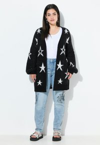 Cardigan en tricot noir avec des motifs d'étoiles blanches, design oversized, porté sur un haut blanc et un jean en denim bleu clair. Des sandales à plateforme noires complètent le look.