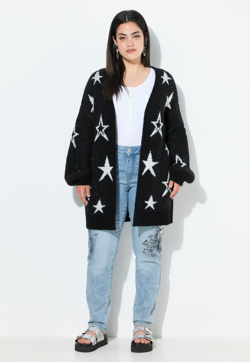 Cardigan en tricot noir avec des motifs d'étoiles blanches, design oversized, porté sur un haut blanc et un jean en denim bleu clair. Des sandales à plateforme noires complètent le look.