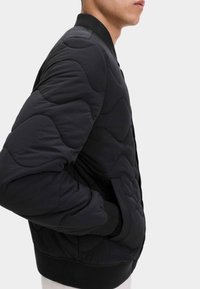 Svart kviltad bomberjacka med ribbad muddar och nederkant. Har en slät yta och rundad, vadderad design. Sidoficka synlig.