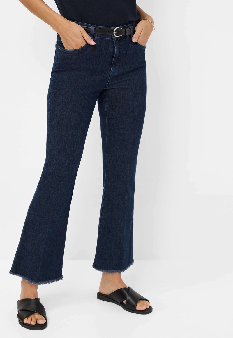 Mørkeblå, flared denim jeans med rå kant forneden, båret med et sort bælte og flade, krydsede sorte sandaler.