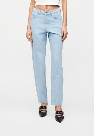 Kvinne iført lyseblå rette jeans og svarte spisse hæler med sølvspenne, stående mot hvit bakgrunn.