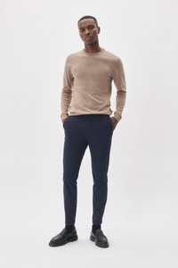 Pull en tricot beige à manches longues associé à un pantalon tailleur bleu marine et des mocassins noirs, sur fond neutre.