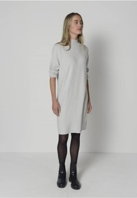 Vestito a maglia grigio chiaro con maniche lunghe, vestibilità comoda, lunghezza a metà coscia e texture a coste. Indossato con stivali neri alla caviglia e collant.