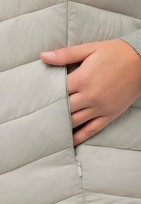 Jasnoszara kamizelka puchowa z haftem w kształcie chevron, z boczną kieszenią zapinaną na zamek i ręką umieszczoną w środku. Gładka tekstura.