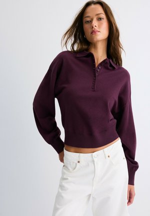 Jeune femme portant un polo en maille violet foncé à manches longues et un pantalon blanc taille haute sur un fond clair uni.