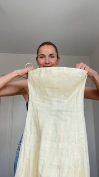 Kremna lanena obleka s strukturiranim dizajnom, ki ima navpične šive in široke naramnice. Lahka tkanina z mehko teksturo.