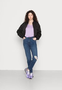 Casaco bomber preto, t-shirt lilás clara, jeans azul de cintura alta com detalhes rasgados e ténis roxos. O modelo está encostado a um fundo liso.