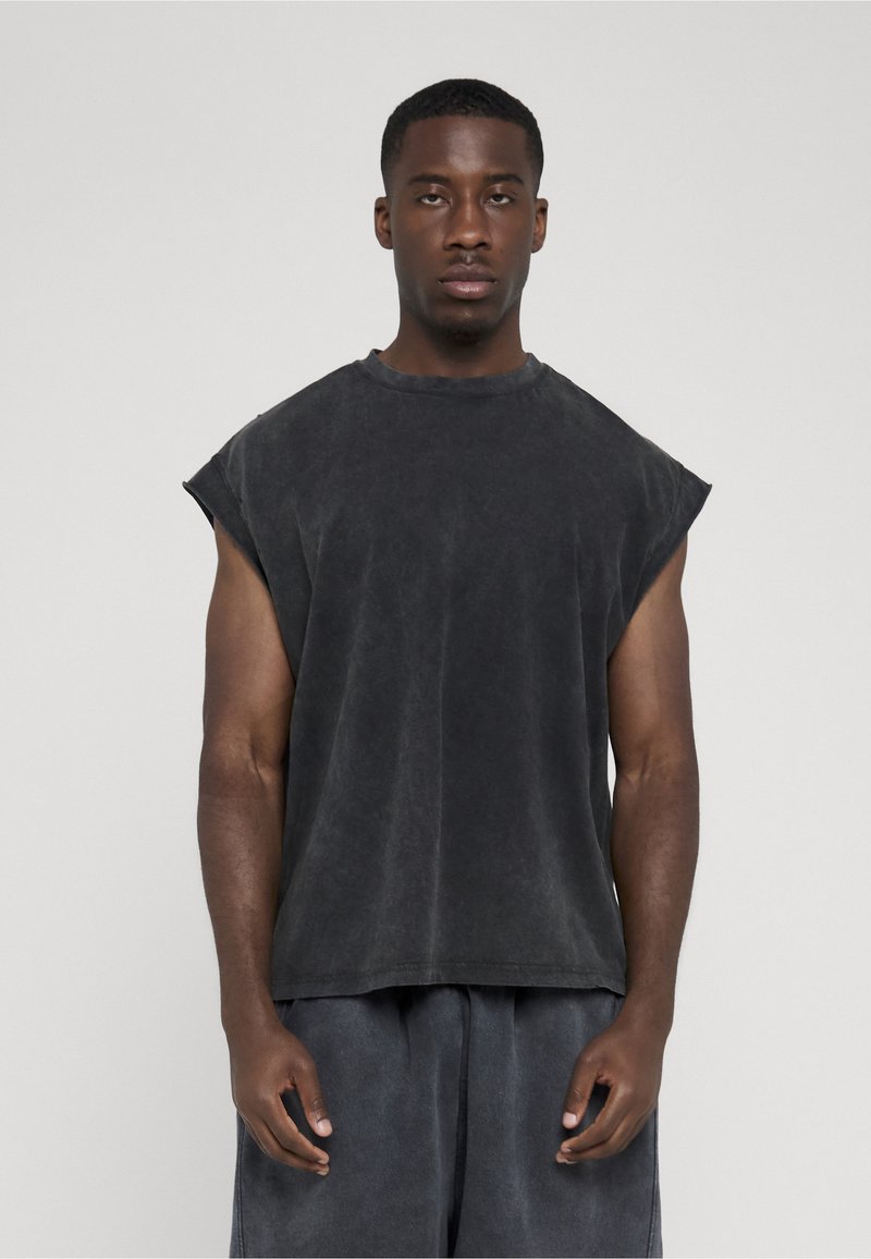 Homme aux cheveux courts portant un t-shirt oversize gris foncé sans manches et un pantalon ample assorti, debout devant un fond clair uni.