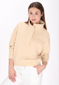Beige pulloveri, jossa on korkea kaulus, napsautusnappikiinnitys ja etutasku. Kangas näyttää pehmeältä, ja helmassa ja hihoissa on ribbireunus. Yhdistetty valkoisiin housuihin.