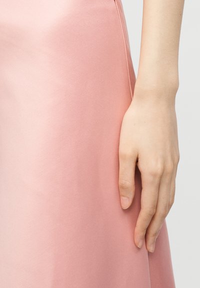 Δεξί χέρι χαλαρό δίπλα σε μια λεία, ανοιχτό ροζ σατέν φούστα πάνω σε ένα απλό φωτεινό φόντο.