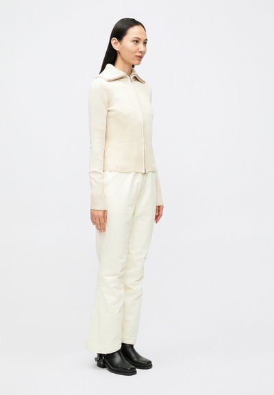 Kremfarget strikket jakke med glidelås og bred krage, ribbet kant, kombinert med hvite bukser og svarte ankelstøvletter med hæl.