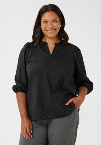 Blouse noire avec un motif texturé, décolleté en V et manches bouffantes. Le tissu semble doux ; associée à un pantalon gris.
