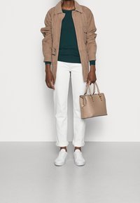 Personne portant une veste beige, un pull vert foncé, un pantalon blanc et des baskets blanches, tenant un sac à main beige sur un fond clair uni.