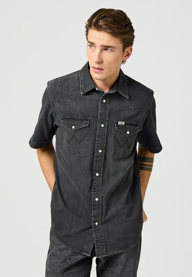 Camicia di jeans a maniche corte grigia scura con cuciture a punta, due tasche sul petto e bottoni bianchi a contrasto. Dettagli di cucitura discreti.