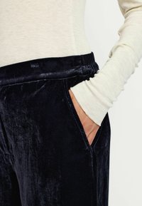 Pantaloni in velluto blu navy con vita elastica e tasche laterali. La texture lucida contrasta con la maglietta a maniche lunghe di colore chiaro.