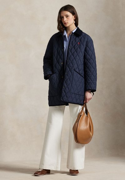 Femeie purtând o haină matlasată bleumarin, cămașă bleu, pantaloni albi cu croială largă, pantofi maro și ținând o geantă de piele maro deschis pe un fundal neutru.