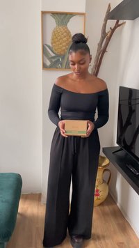 Μαύρη μπλούζα με μακριά μανίκια και χωρίς ώμους συνδυασμένη με φαρδιά μαύρα παντελόνια; κρατώντας ένα μπεζ κουτί με πράσινες λεπτομέρειες. Τέχνη ανανά στο φόντο.