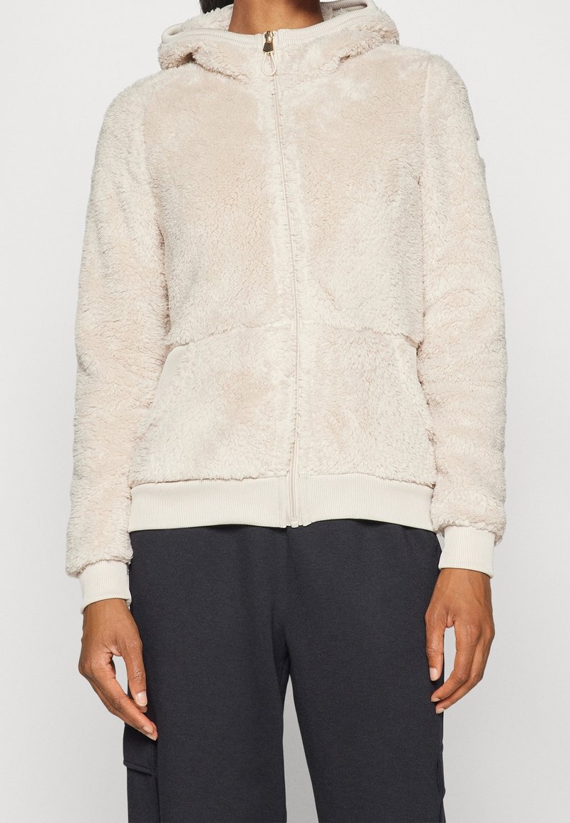 Personne portant un sweat à capuche zippé beige clair, duveteux, avec des poignets et un ourlet côtelés, assorti à un pantalon foncé sur un fond uni.