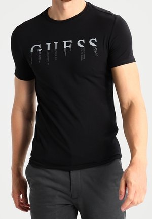 Mężczyzna w dopasowanej czarnej koszulce z krótkim rękawem z logo "GUESS" w srebrnych literach na piersi, w połączeniu z ciemnoszarymi spodniami.