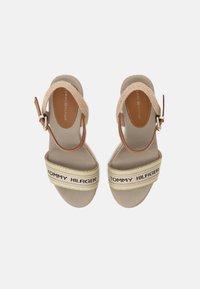 Brune sandaler med en kraftig sål, der har en bred rem med "TOMMY HILFIGER" i sort, accentueret af gule og beige striber. Justerbar ankelrem.