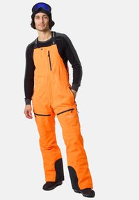 Heldere oranje skibroeken met zwarte accenten, voorzien van ritszakken en verstelbare banden, ontworpen voor wintersport en buitenactiviteiten.