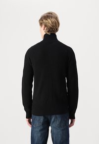 Svart ribbestrikket genser med høy krage og lange ermer, kombinert med blå jeans med baklommesøm i sikksakkmønster.