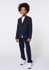 Completo blu navy con una vestibilità slim, caratterizzato da due bottoni e un risvolto a scollo, abbinato a una camicia bianca e scarpe da ginnastica bianche.