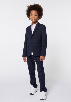 Completo blu navy con una vestibilità slim, caratterizzato da due bottoni e un risvolto a scollo, abbinato a una camicia bianca e scarpe da ginnastica bianche.