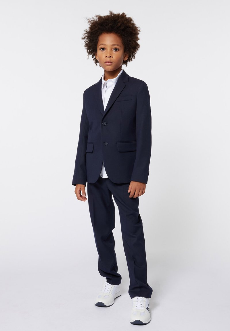 Completo blu navy con una vestibilità slim, caratterizzato da due bottoni e un risvolto a scollo, abbinato a una camicia bianca e scarpe da ginnastica bianche.