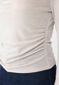 Jasnobeżowa bluzka z długim rękawem o delikatnym połysku, z marszczeniami po bokach dodającymi tekstury oraz dopasowanym krojem. Połączona z ciemnymi dżinsami.