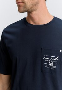 Mörkblå bomull t-shirt med korta ärmar, rund halsringning och en vänster bröstficka med vit tryckt text och en tryckknapp.