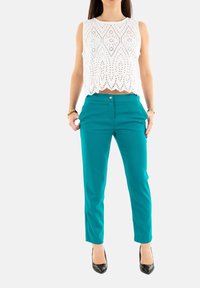 Haut court en dentelle blanche avec un ourlet festonné, associé à un pantalon slim-fit teal avec des poches avant et une fermeture à bouton. Des talons noirs viennent parfaire le look.