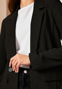 Černý žebrovaný blazer s klopovým límcem, se zapínáním na jeden knoflík a bílým tričkem pod ním; ruka umístěna na blazru.
