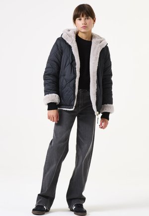 Veste noire matelassée avec une capuche et un ourlet en fausse fourrure grise, portée sur un pull noir et un jean gris, dotée d'une fermeture éclair et de poches latérales.