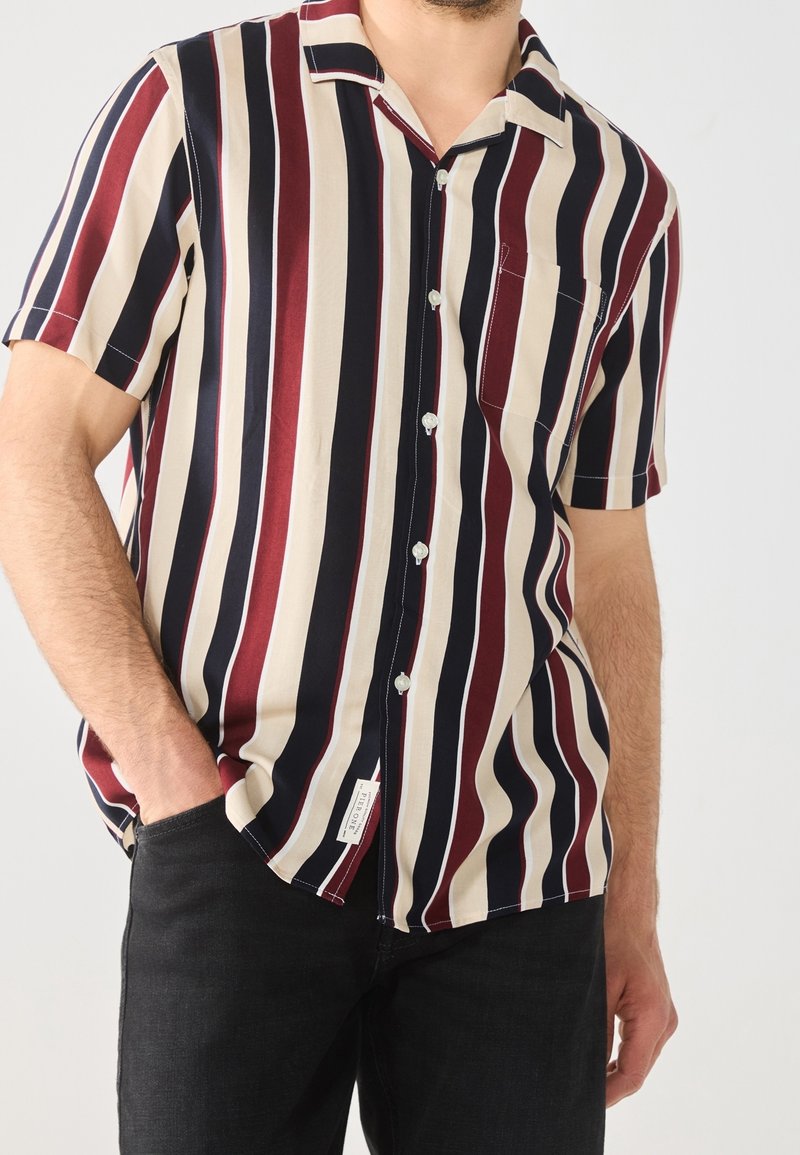 Korte mouwen shirt met verticale strepen in bordeauxrood, marineblauw en crème. Heeft een knoopsluiting aan de voorkant en een enkele borstzak. Zachte stof.