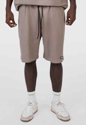 Hombre vistiendo pantalones cortos texturizados color topo con cordones negros, calcetines blancos con texto y zapatillas blancas con detalles en beige y gris.