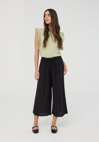 Top verde chiaro con maniche in pizzo arricciato, abbinato a pantaloni neri a gamba larga e ballerine nere. Tessuti lisci, vestibilità rilassata.
