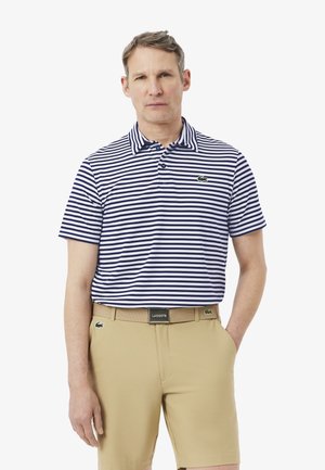 Homme portant un polo rayé bleu marine et blanc et un short beige avec une ceinture assortie, debout avec une main dans la poche.