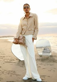 Vrouw in een beige houndstooth jasje, witte wijde broek, witte schoenen, zonnebril, met een beige gewatteerde handtas op een zandstrand.