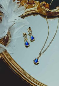 Collier en or et boucles d'oreilles assorties ornés de pierres en forme de goutte d'eau bleues, entourées de pierres transparentes, déposés sur une surface miroir avec des plumes.