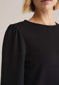 Nő texturált fekete, hosszú ujjú felsőt visel, enyhén fodros vállakkal és kerek nyakkivágással, egyszerű bézs háttér előtt.