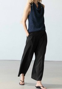 Femme portant un top sans manches bleu marine à col bénitier, un pantalon noir à jambes larges et des tongs noires, debout les mains dans les poches.