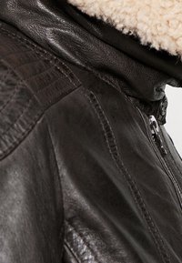 Giacca di pelle marrone con superficie testurizzata. Presenta un colletto in shearling, una cerniera metallica e cuciture diagonali sulla spalla.