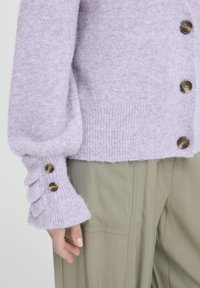 Lila gestrickte Strickjacke aus weichem Stoff, mit gerippten Bündchen und Knopfdetails sowie einer lockeren Passform. Kombiniert mit hellgrünen Hosen.