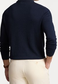 Pull en maille bleu marine avec col et manches longues, présentant une texture côtelée et une coupe ajustée, associé à un pantalon de couleur crème.
