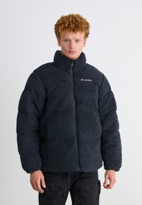 Zwarte, met fleece gevoerde pufferjack met hoge kraag, ritssluiting en zijzakken. Heeft een gestructureerde, pluizige buitenkant en gestikte details.