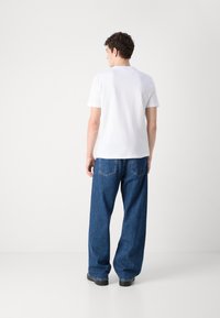 Hvid kortærmet t-shirt parret med løse blå denim jeans med synlige baglommer, sat på en ensfarvet lys baggrund.
