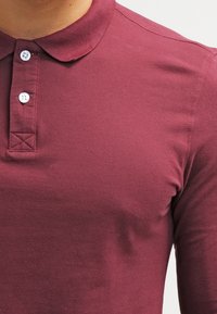 Polo de color burdeos con un acabado texturizado, que presenta un collar, tapeta de tres botones y detalles de punto cruzado por encima de los botones.
