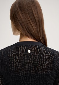 Top nero trasparente con un motivo reptile testurizzato, rifinito con un accento in metallo sul collo dietro. Tessuto liscio e leggero.