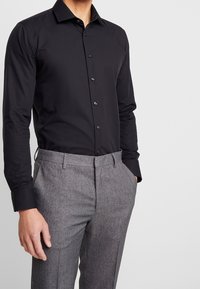 Man som bär en åtsittande svart skjorta med knappar instoppad i grå texturerade kostymbyxor med händerna i fickorna, mot en enkel ljus bakgrund.